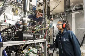 Two students with ear protectors and protective eye glasses look at a big complicated machine